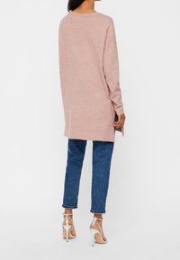Frau in einem langen, staubrosa Pullover, blauen, cropped Jeans und weißen hohen Sandalen, die mit dem Rücken zu einer weißen Wand steht.