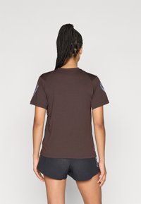 Brązowa sportowa koszulka z krótkim rękawem, o zaokrąglonym dekolcie i kontrastujących ciemnych panelach na ramionach. Na rękawach subtelne oznakowanie marki.