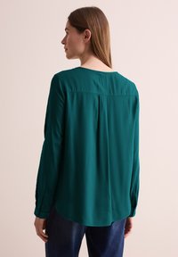 Blusa lunga teal a manica lunga con scollo rotondo, dettagli con pieghe sul retro e polsini bottonati, realizzata in un tessuto liscio e testurizzato.
