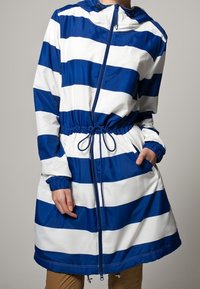 Blå och vit randig vattentålig jacka med huva, åtsittande midja, framfickor och elastiska ärmslut. Slät textur, avslappnad passform.