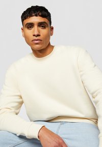 Lys beige genser laget av mykt stoff, med rund halslinning og ribbede mansjetter. Modellen poserer uformelt, iført lyseblå jeans.