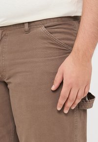 Brown cotton trousers with a textured finish, featuring a side pocket and a slim fit, shown with a hand resting on the thigh area.