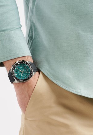 Montre avec bracelet en caoutchouc noir, cadran teal, lunette argentée et index des heures contrastants. Portée sur une chemise teal clair et un pantalon kaki.