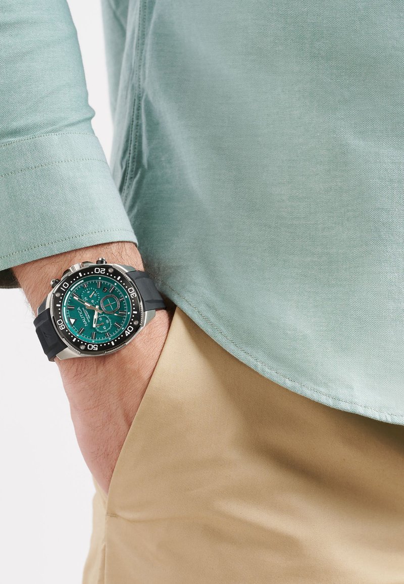 Orologio con cinturino in gomma nera, quadrante color teal, lunetta argentata e indici delle ore in contrasto. Indossato su una camicia di un leggero color teal e pantaloni khaki.