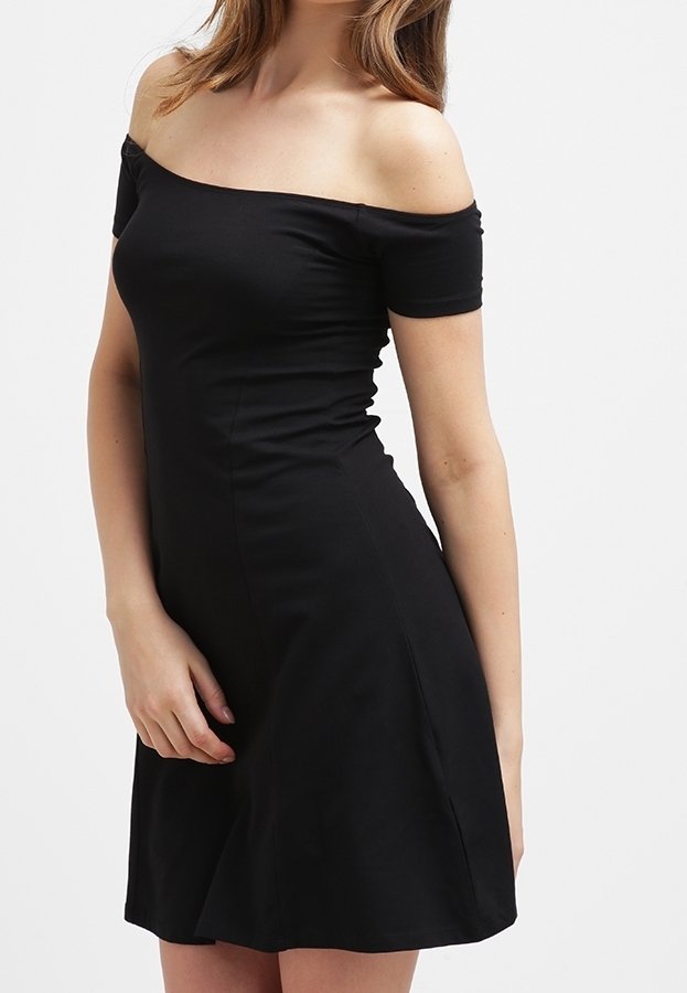 Femme portant une robe noire ajustée, épaules dénudées, manches courtes, debout devant un fond clair uni.