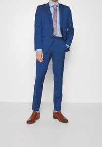 Niebieski garnitur o slim fit, marynarka z jednym rzędem guzików, klapy notch oraz dopasowane spodnie. Brązowe skórzane buty i wąski krawat w paski dopełniają stylizację.