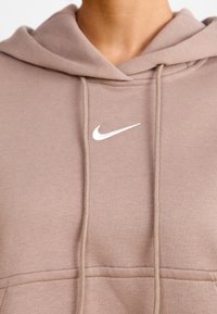 Närbild av en person som bär en beige huvtröja med en vit Nike-logotyp och snörning vid bröstområdet.
