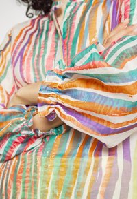 Personne aux bras croisés portant un chemisier coloré à manches longues avec des rayures verticales orange, violettes, vertes, bleues et blanches.