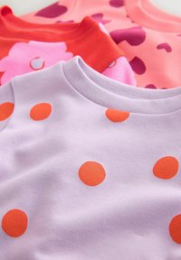 Trois hauts à manches courtes : lavande avec des pois orange, rose avec des cœurs foncés et rouge avec des fleurs roses ; fabriqués en tissu doux, design à col rond.