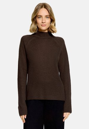 Femme aux cheveux blonds portant un pull en tricot marron foncé à manches longues et un pantalon noir, debout devant un fond clair uni.