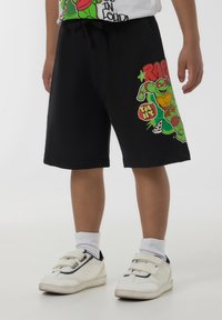 Pantaloni corti neri in misto cotone con una grafica che presenta un personaggio verde e del testo. La vita include un cordino per la regolazione.
