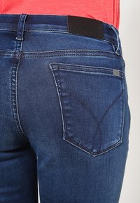 Jeans de mezclilla azul oscuro con un diseño ajustado, costuras prominentes y una forma distintiva de los bolsillos traseros que presenta un logo sutil.