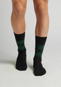 Ben som bär svarta ankelsockar med grön och mörk mönstrad argyle-design, står på ett ljusgrått golv mot en enkel bakgrund.