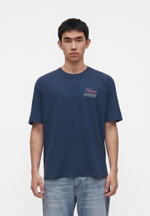 Jeune homme aux cheveux noirs courts portant un t-shirt bleu marine Miami Williams Racing et un jean bleu clair, debout sur un fond blanc.