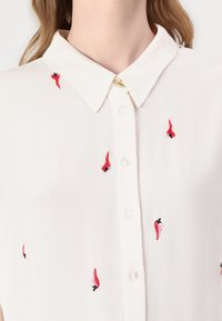 White blouse with a collar, featuring red chili pepper embroidery. Designed with a button front and soft, lightweight fabric.