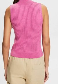 Henkilö, joka on pukeutunut hihattomaan, ribbaiseen vaaleanpunaiseen toppiin ja vaaleisiin housuihin, joissa on takatasku, kuvattuna taustalta yksinkertaisen taustan edessä.