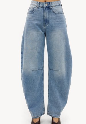Jean en denim bleu clair à jambes larges, taille haute avec des détails de couture visibles aux genoux, porté avec des chaussures pointues marron foncé.