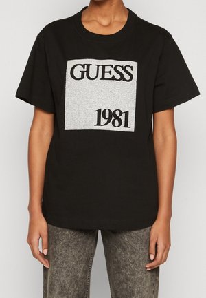 Czarna koszulka z krótkim rękawem z srebrnym kwadratem z napisem "GUESS" i "1981" czarnym tekstem, noszona z szarymi jeansami typu acid-wash.