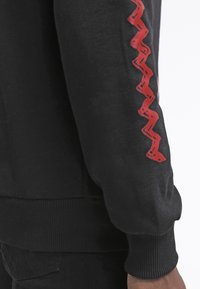 Felpa nera con tessuto testurizzato, polsini a coste e un distintivo motivo a zigzag rosso sulla manica sinistra.