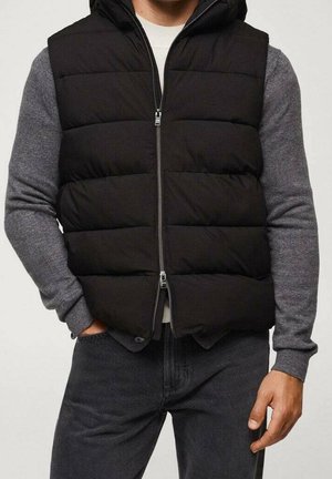 Mand iført en sort quiltet ærmeløs vest over en grå langærmet sweater, mørke jeans, med den ene hånd i lommen og den anden synlig.