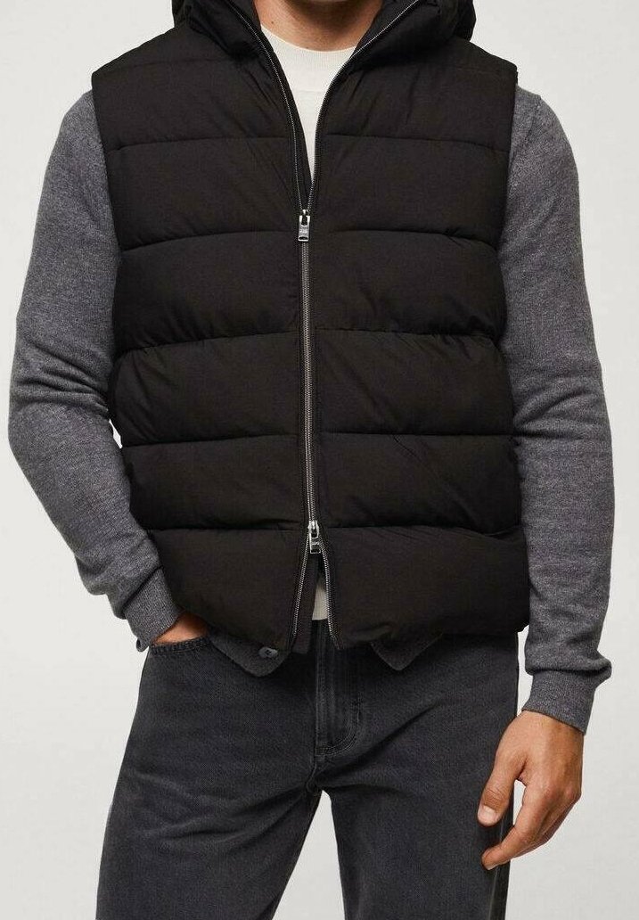 Homme portant une veste sans manches matelassée noire sur un pull gris à manches longues, jean foncé, avec une main dans la poche et l'autre visible.