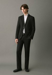 Completo nero con giacca su misura e pantaloni coordinati, abbinato a una camicia bianca. Tessuto liscio, vestibilità slim, design minimalista.