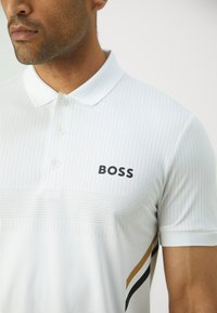 White polo shirt with a textured pattern, featuring a black "BOSS" logo, short sleeves, and contrasting gold and black stripes on the side.