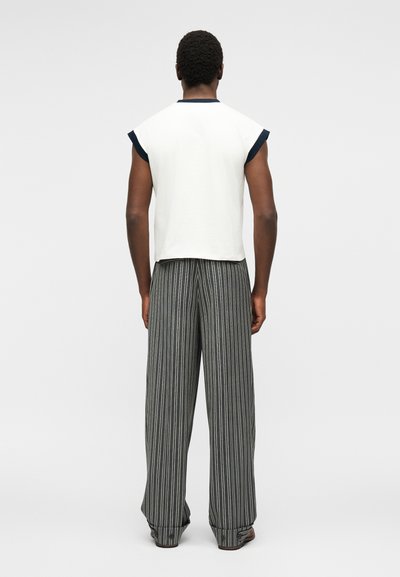Άνδρας που στέκεται με την πλάτη, φοράει άσπρο αμάνικο τοπ με μαύρη επένδυση και ανοιχτόχρωμο γκρι παντελόνι με ρίγες.