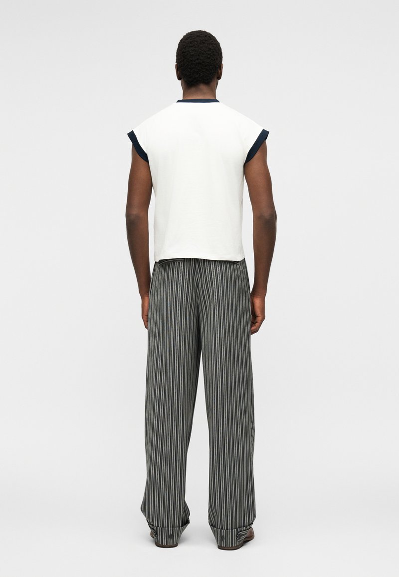 Man standing facing away wearing white sleeveless top with black trim and loose gray pinstriped pants.