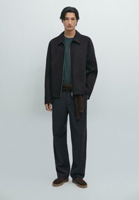 Zwarte ritsjack over een donkergroene shirt, relaxte, donkere broek en bruin suede schoenen. Eenvoudig ontwerp zonder zichtbare patronen.