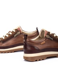 Zapatillas de piel marrón con cordones y acentos de color crema. Cuentan con un cierre de cremallera y una suela de goma resistente. Tacón marrón texturizado y entresuela beige.