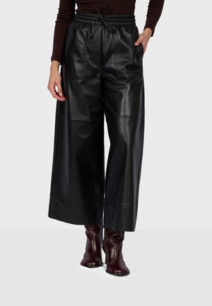 Personne portant un pantalon en cuir noir à jambes larges avec une taille à cordon et des bottines marron foncé, debout devant un fond uni.