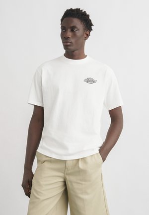 Νέος άνδρας που φορά λευκό κοντομάνικο μπλουζάκι με μικρό σχέδιο στο στήθος και μπεζ παντελόνι, στέκεται με το ένα χέρι στην τσέπη, κοιτάζοντας πλάγια.