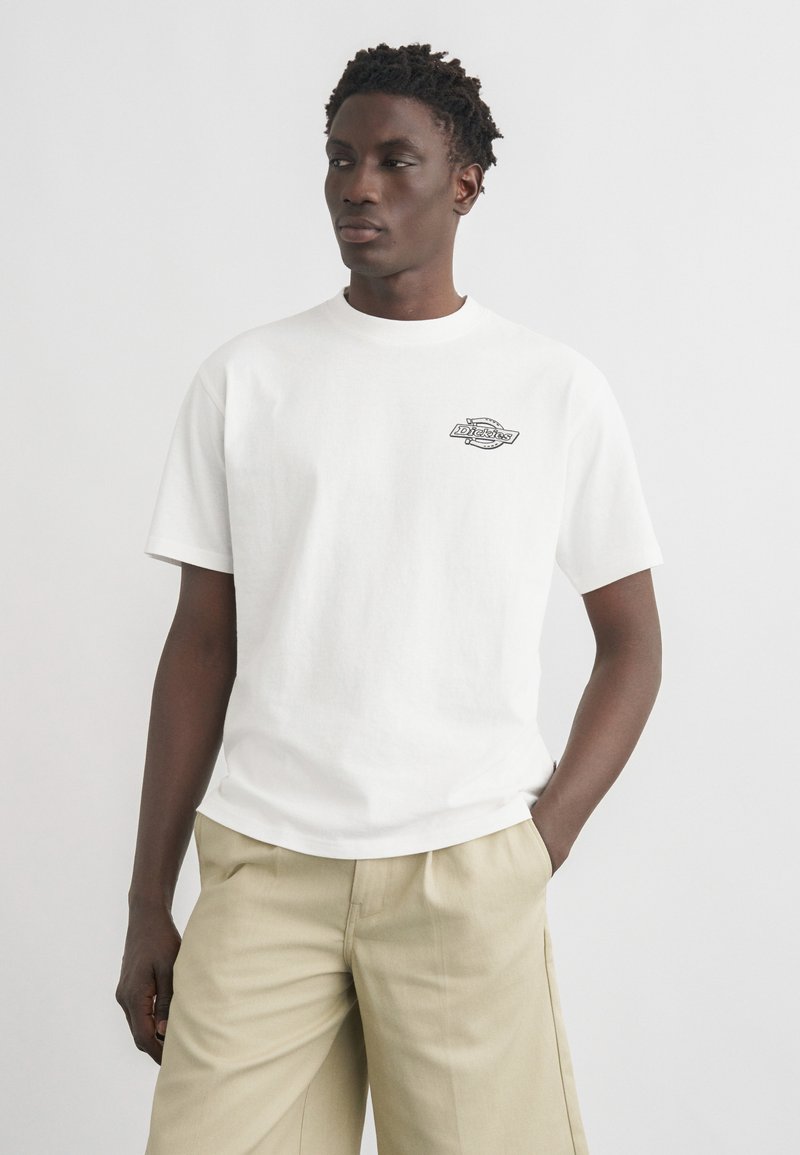 Jeune homme portant un t-shirt blanc à manches courtes avec un petit motif sur la poitrine et un pantalon beige, debout avec une main dans la poche, regardant sur le côté.