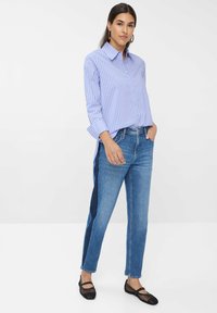 Donna in piedi con una mano in tasca, che indossa una camicia a righe blu, jeans blu bicolore e ballerine nere in rete, su sfondo bianco.