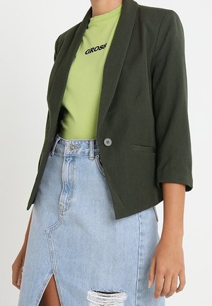 Kvinde iført en mørkegrøn blazer over en lysegrøn "GROSS" t-shirt og en lyseblå denimnederdel med en frontslids og slidte detaljer.