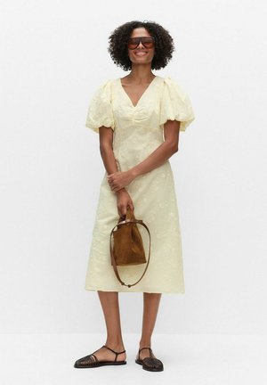 Femme aux cheveux bouclés portant de grandes lunettes de soleil, une robe jaune clair à manches bouffantes, des sandales noires, et tenant un sac à main en daim marron.