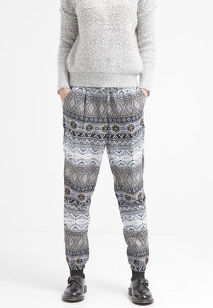 Person trägt grau texturierten Pullover, gemusterte lockere Hose mit geometrischen Mustern und schwarze Schnallenschuhe, Hände in den Taschen, stehend auf weißem Hintergrund.