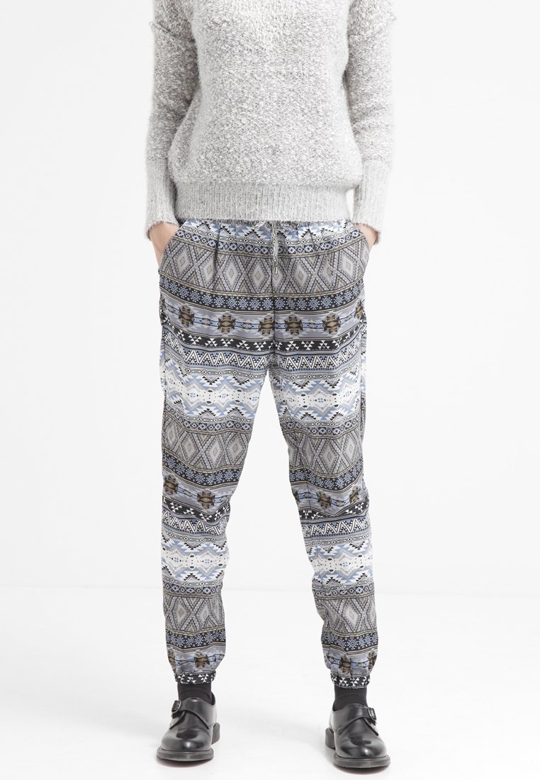 Personne portant un pull gris texturé, un pantalon ample à motifs géométriques et des chaussures noires à boucle, mains dans les poches, debout sur fond blanc.