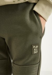Grüne Jogginghose mit weicher Textur, mit einem Aufnäher, auf dem in weißer Schrift "PLAY ALL DAY" steht, und gerippten Bündchen an den Knöcheln.
