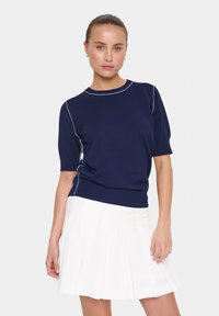 Pull bleu marine à manches courtes avec un col rond, présentant des accents de couture blanche le long des manches et du corps, associé à une jupe plissée blanche.