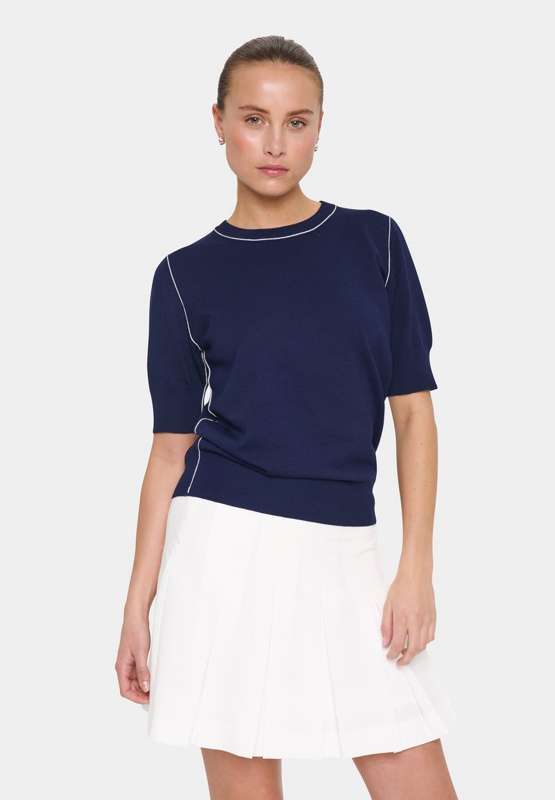 Pull bleu marine à manches courtes avec un col rond, présentant des accents de couture blanche le long des manches et du corps, associé à une jupe plissée blanche.