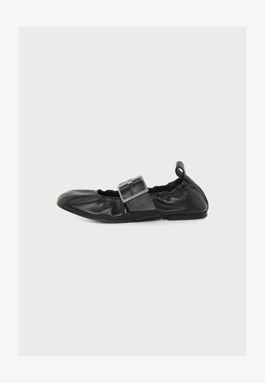 Zapato plano de cuero negro con una gran hebilla plateada y laterales elásticos sobre fondo blanco liso.