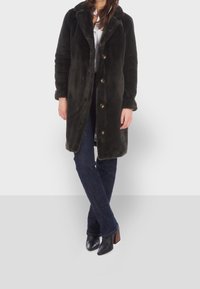 Mörkgrön konstpälsjacka med slagkrage, prydd med guldknappar, sidofickor och en mjuk yta, buren över mörka jeans och svarta klackar.