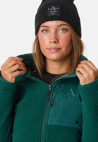 Giacca in pile verde con zip frontale, caratterizzata da una texture morbida, logo ricamato e una tasca nera a toppa. Berretto di maglia nero sulla testa.