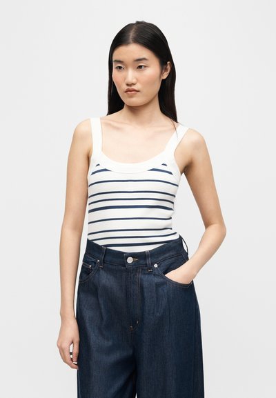 Young woman with long dark hair wearing a white tank top with navy horizontal stripes and high-waisted wide-leg dark blue jeans.
