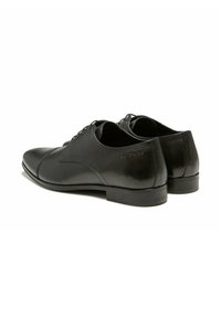 Chaussures habillées en cuir noir présentant un design élégant, un bout pointu, une texture lisse et un talon plat. Nom de la marque embossé sur le côté.