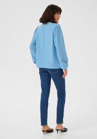 Haut long à manches longues bleu clair texturé avec des manches bouffantes, associé à un jean slim bleu foncé et des mules à talons noires.