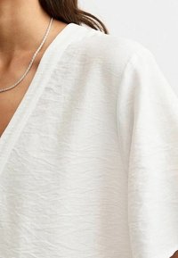 Bílý texturovaný top s hlubokým výstřihem do V a širokými rukávy. Je vidět stříbrný řetízek, který dodává jemný detail.