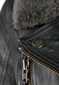 Zwarte leren jas met een grijze bontvoering in de kraag, voorzien van een metalen drukknoop en gouden ritsdetails aan de voorkant en de zak.